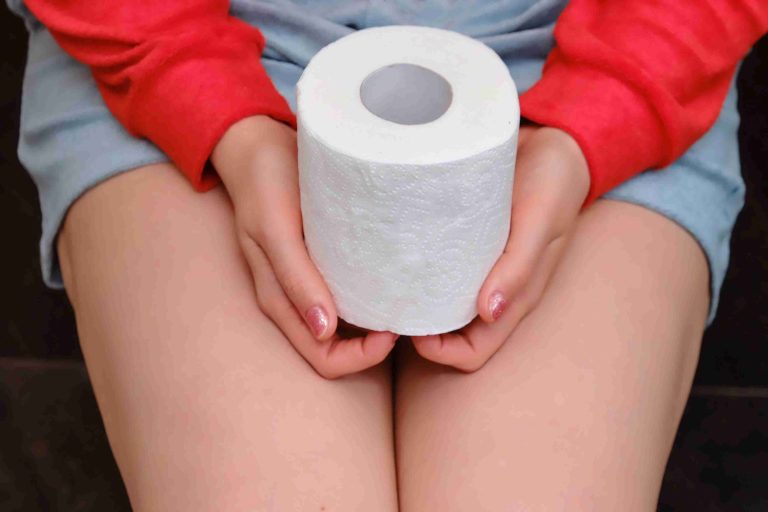 Woman sitting on the toilet bowl holding a roll of toilet paper.