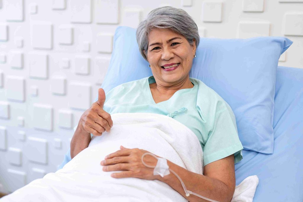 Elderly patient smiling and giving the thumbs up while lying in a hospital bed.