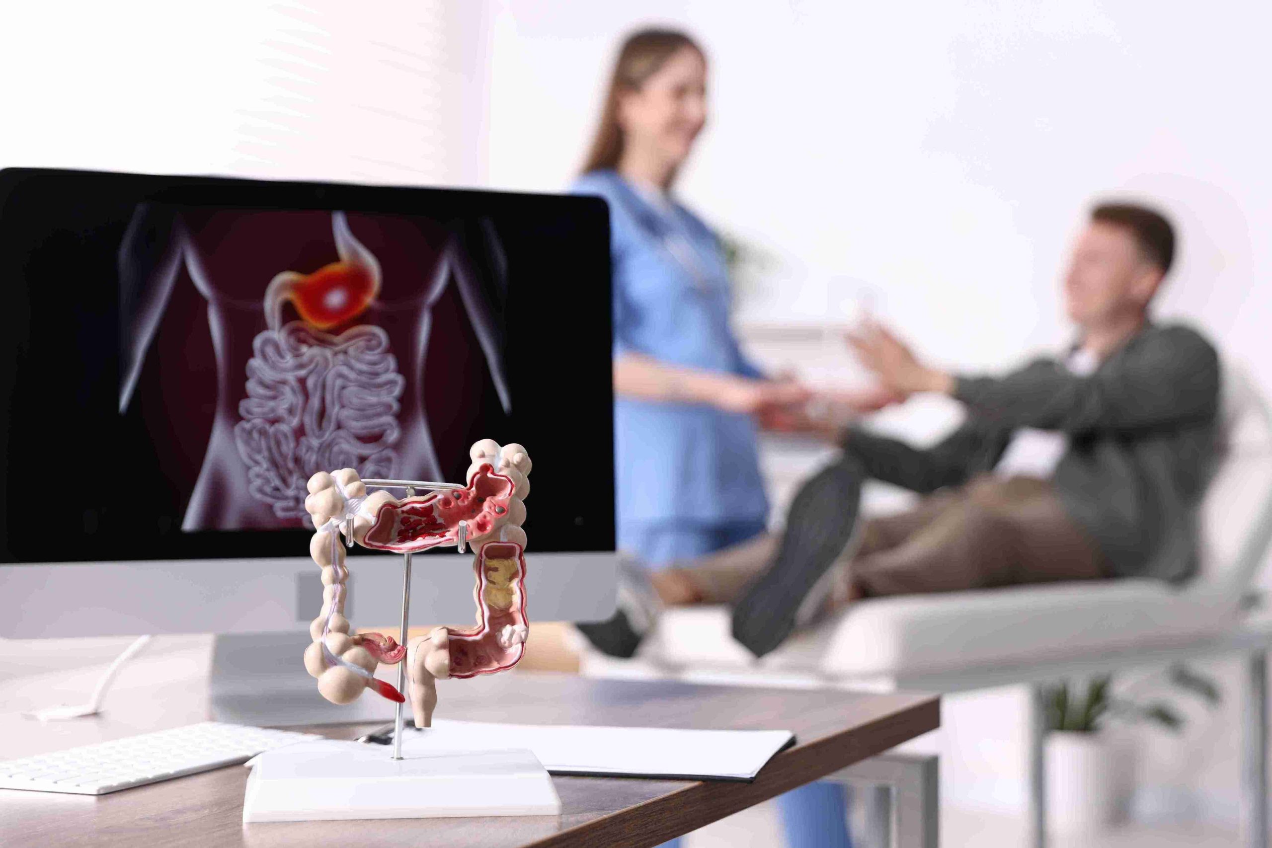Image of an anatomical colon model with patient and healthcare worker in the background.